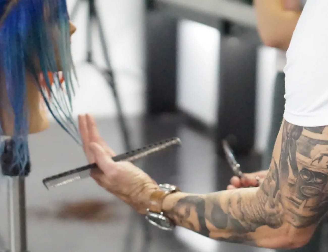 Hairstylist with tattoos trims blue hair on a mannequin.