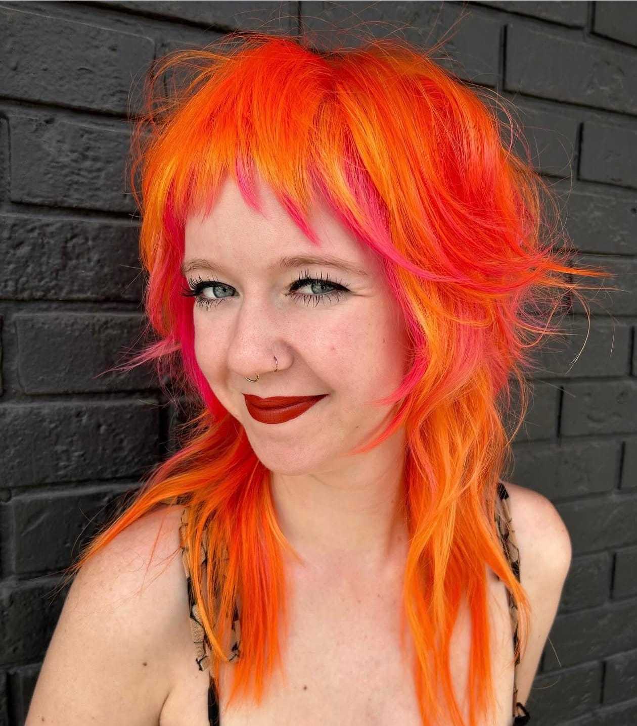 Young woman with vibrant orange and pink hair smiling against a black brick wall.