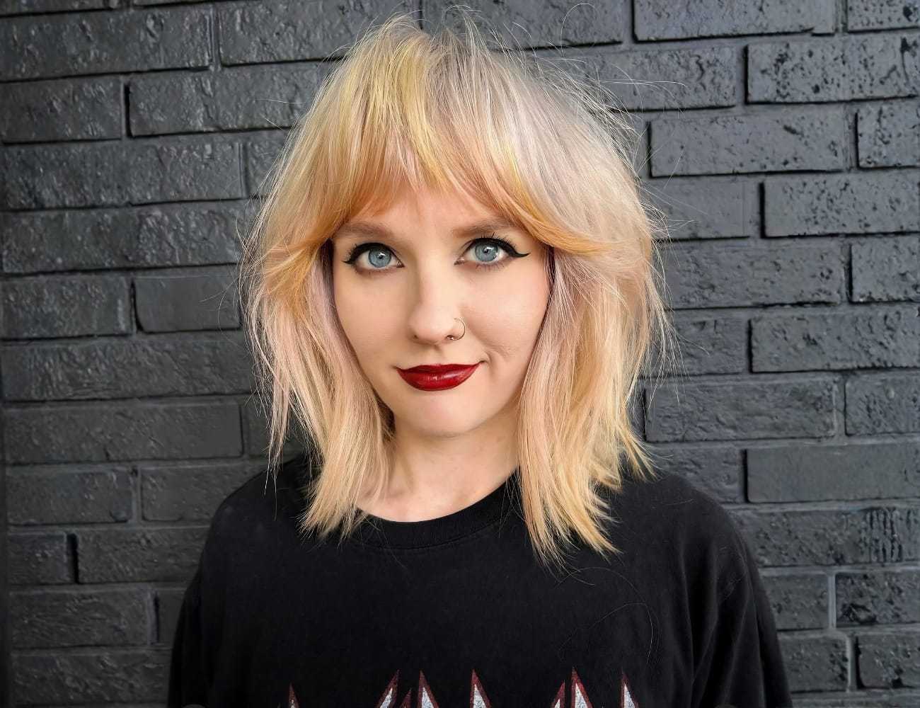 Young woman with blonde hair and striking makeup stands against a dark brick wall.