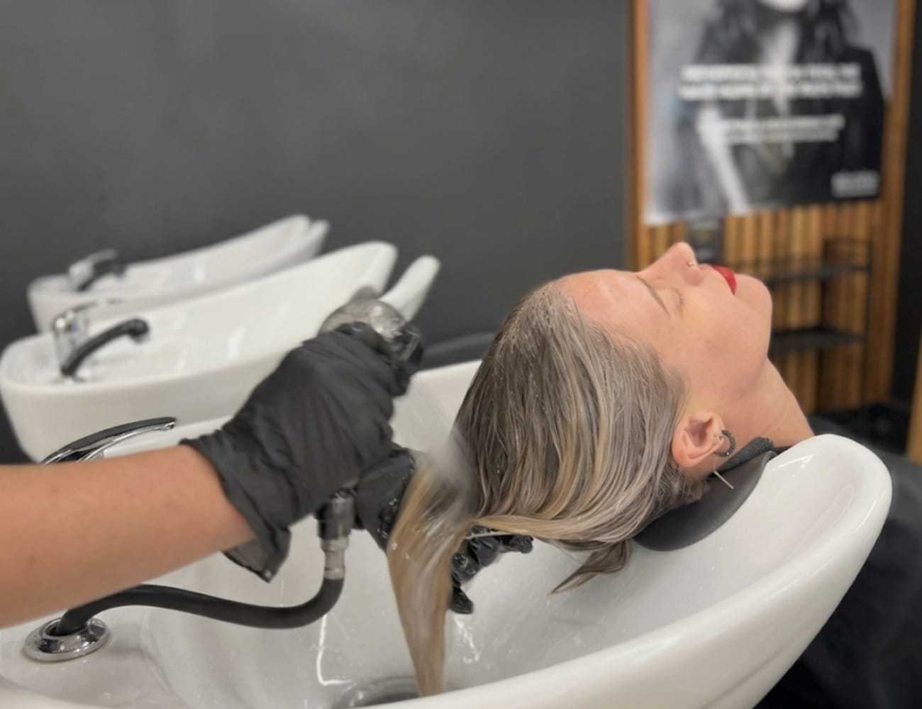 Hair stylist washing a woman's hair in a salon sink, with black gloves and reclining chair.